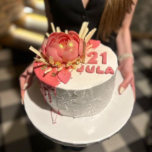 Osoba trzyma tort urodzinowy z białą polewą, różowym kwiatem z lukru, napisem '21 Jula' i świeczką.