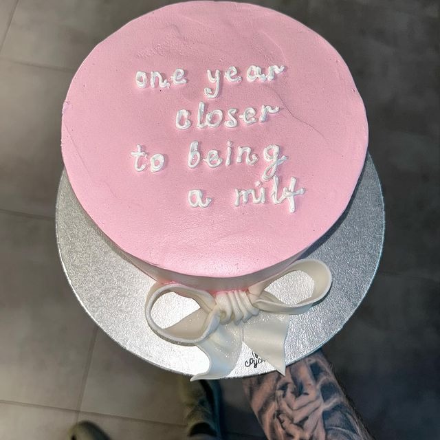 Różowy tort urodzinowy z ironicznym napisem 'one year closer to being a milf', udekorowany białym lukrowym kokardą, na srebrnej podkładce