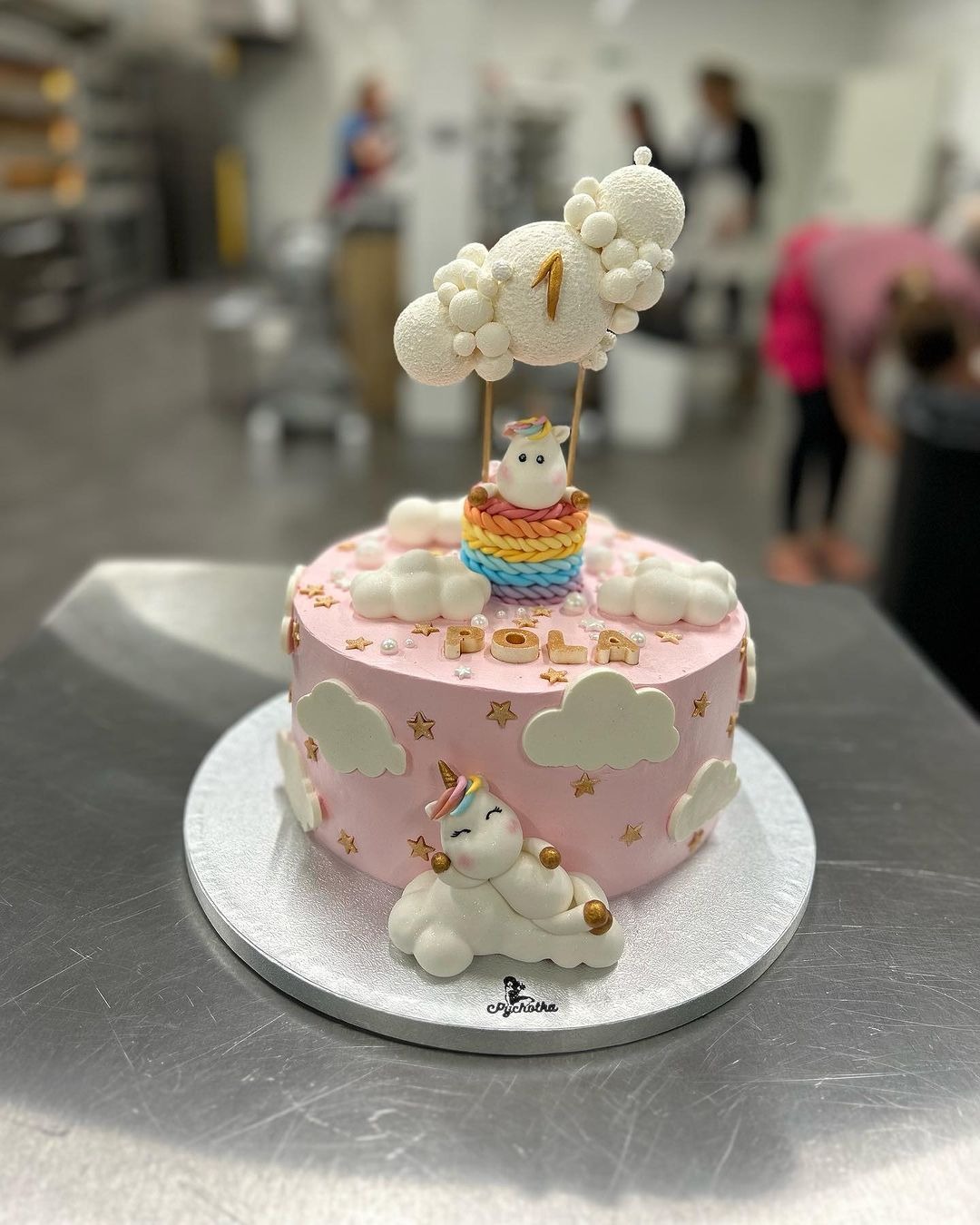 Różowy tort urodzinowy z motywem jednorożca i chmur, ozdobiony napisem 'POLA' i topperem w kształcie chmury z cyfrą '1', na metalowym stole, w tle profesjonalna kuchnia cukiernicza.