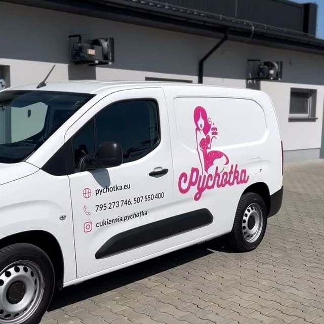 Biały samochód dostawczy z różowym logo i danymi kontaktowymi firmy cukierniczej 'Pychotka' na boku, zaparkowany na zewnątrz budynku.