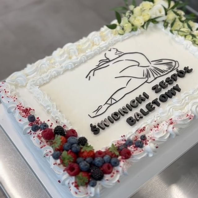 Prostokątny tort z białym kremem, dekorowany owocami leśnymi i kwiatami. Na górze tortu czarny rysunek baletnicy i napis 'Świdnicki Zespół Baletowy' wykonany z czarnych literek.