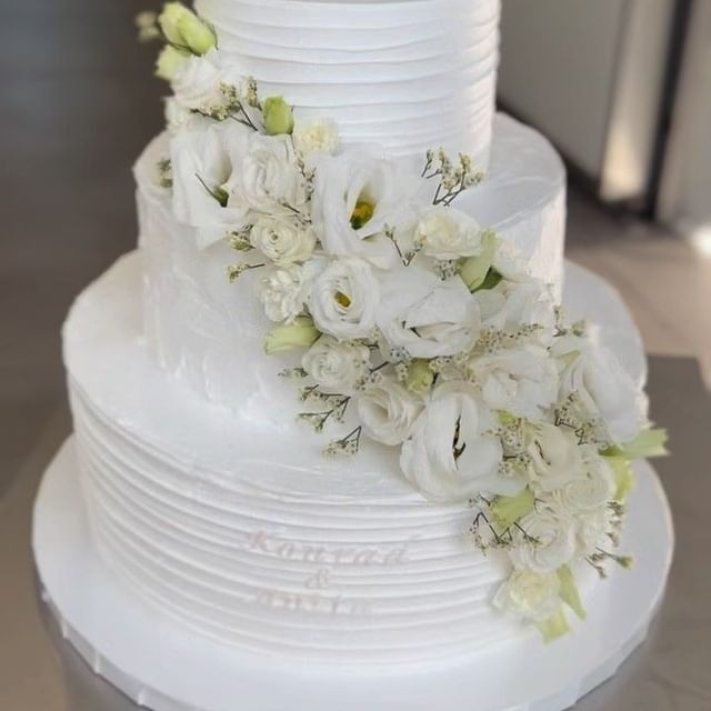 Elegancki, trzypiętrowy tort weselny udekorowany białymi kwiatami, z delikatnym teksturowaniem kremu i subtelnym napisem z imionami na dolnym poziomie.