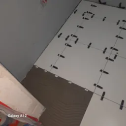 Białe płytki podłogowe w trakcie układania, z użyciem krzyżyków dystansowych i poziomowania, widoczna warstwa kleju na podłożu w rogu pomieszczenia.