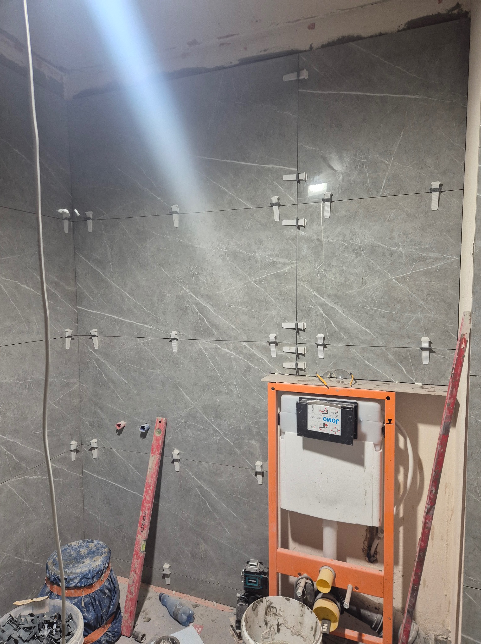 Ściana łazienki wyłożona szarymi płytkami imitującymi marmur, z użyciem klipsów poziomujących, widoczny stelaż podtynkowy WC z zamontowaną spłuczką, wiadro z zaprawą i poziomica.