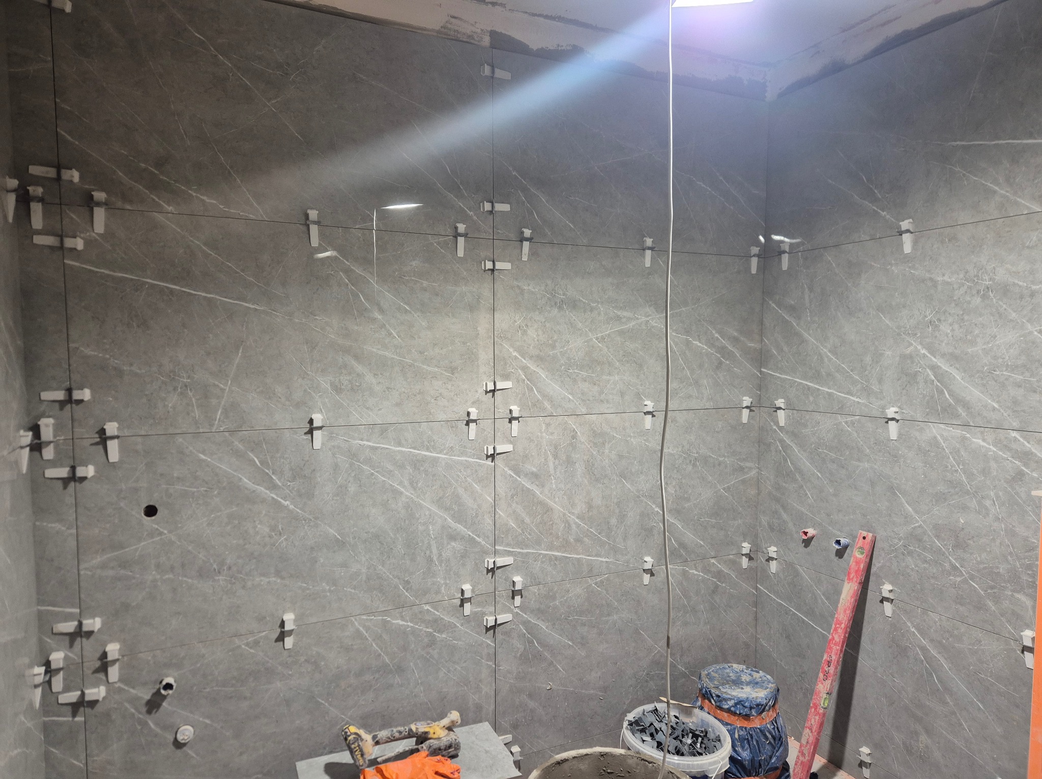 Świeżo ułożone, szare płytki imitujące marmur na ścianach łazienki, z białymi krzyżykami dystansowymi, poziomica oparta o ścianę, wiaderka i narzędzia glazurnicze w rogu.