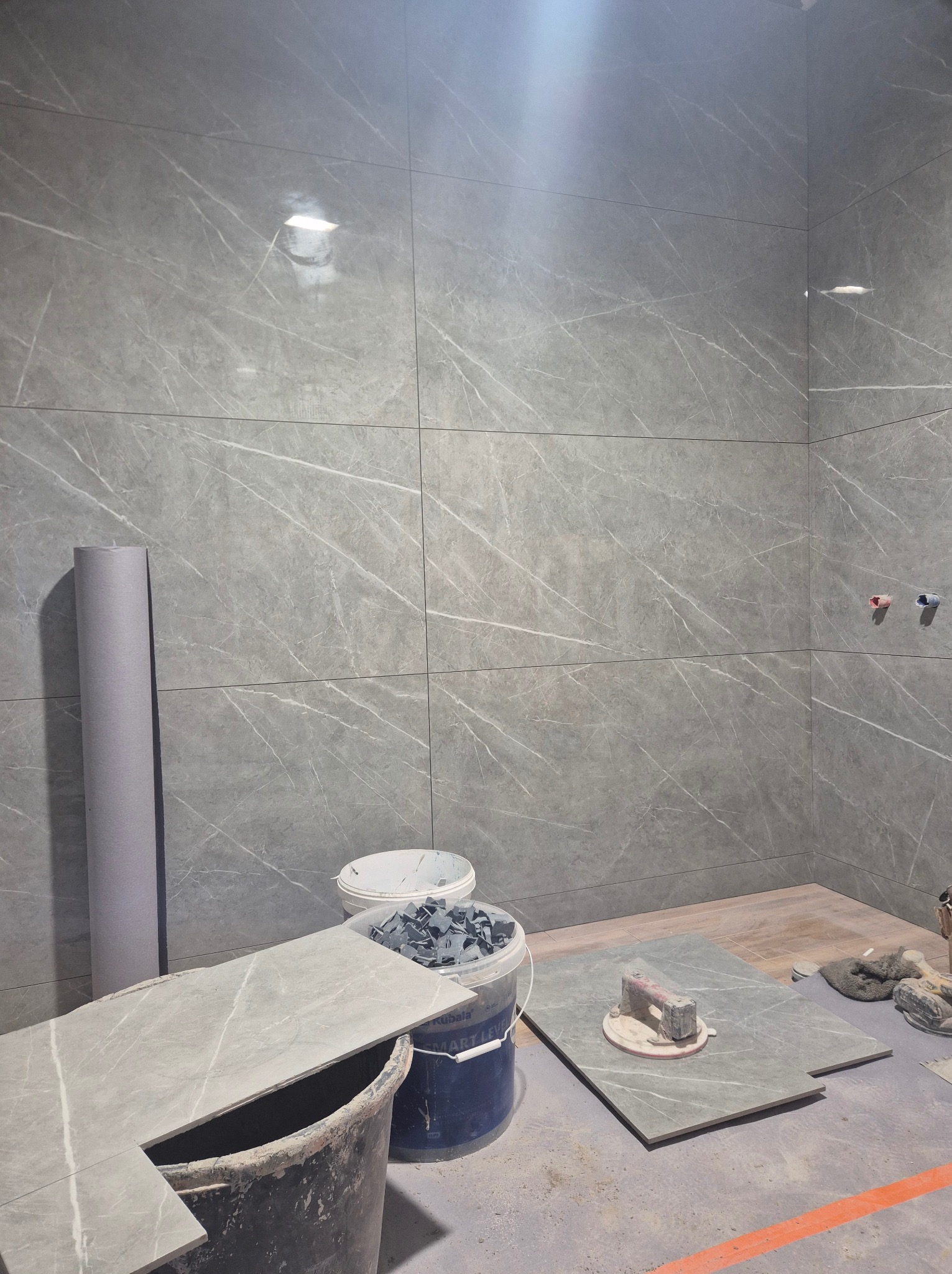 Ściana łazienki w trakcie układania płytek imitujących marmur, widoczne narzędzia glazurnicze, wiadro z klejem, rolka folii ochronnej oraz niebieskie i czerwone zaślepki na rurach.