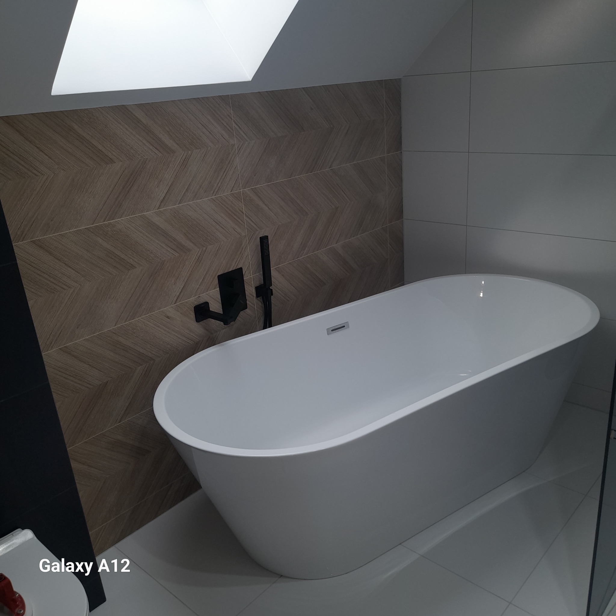 Nowoczesna, wolnostojąca wanna owalna z czarną armaturą na tle ściany z płytek imitujących drewno ułożonych w jodełkę. Widoczny skos sufitu z oknem.