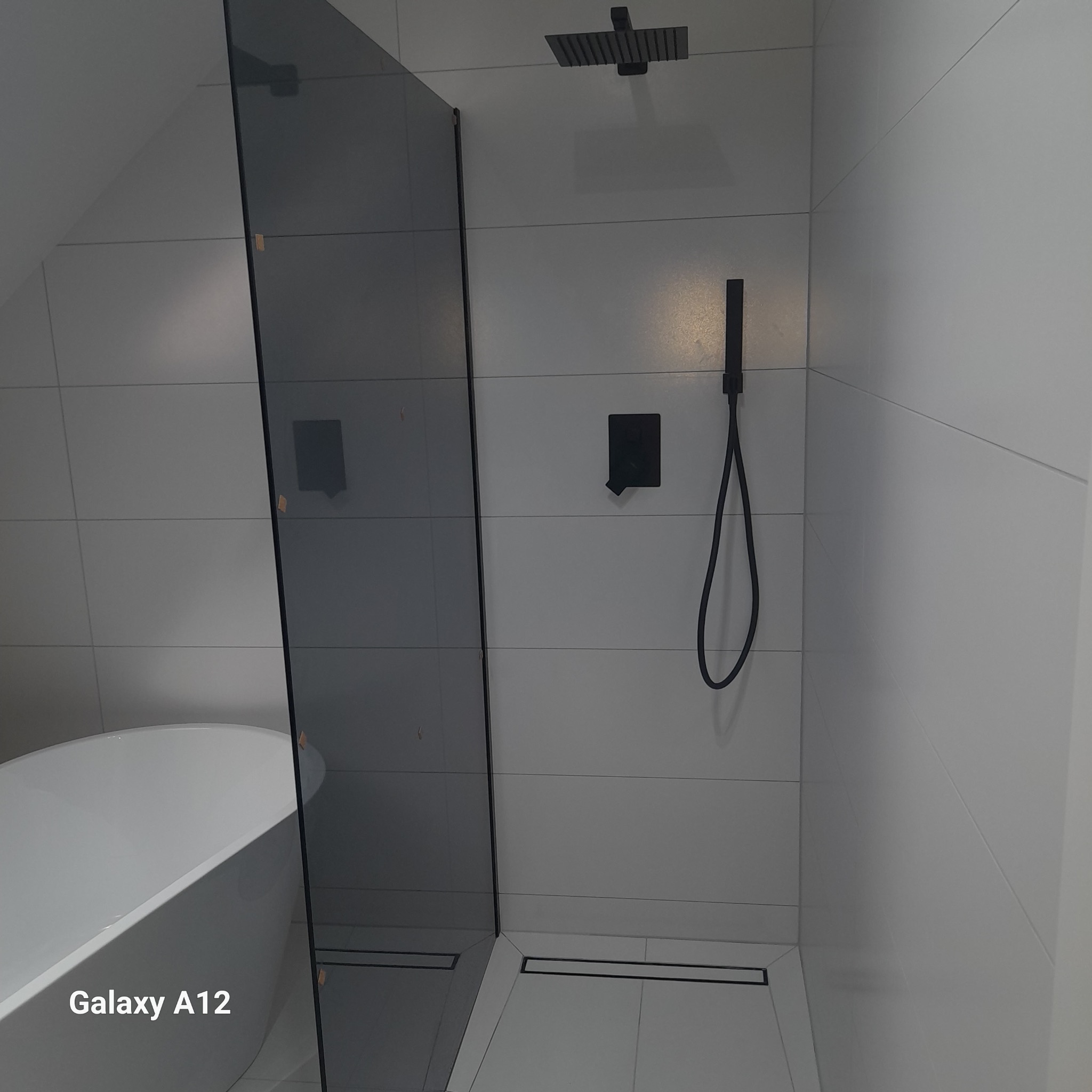 Nowoczesna łazienka w odcieniach szarości z kabiną prysznicową typu walk-in z grafitową szybą, deszczownicą i zestawem natryskowym w czarnym macie oraz wanną wolnostojącą.