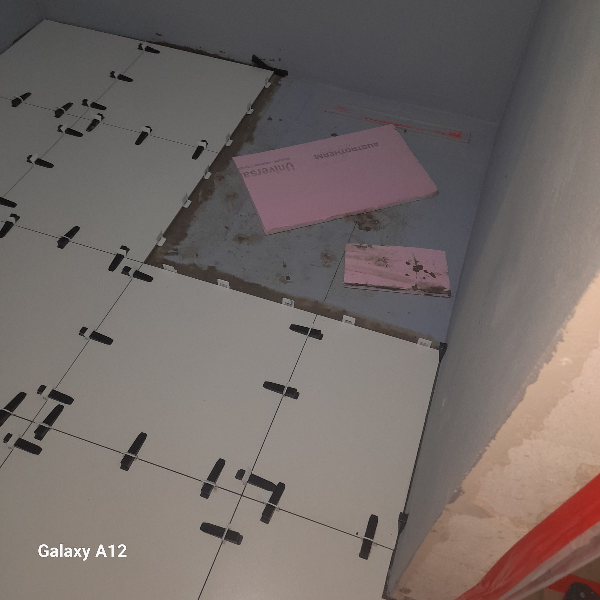 Układanie białych płytek z czarnymi krzyżykami dystansowymi w trakcie prac w Bielsko-Białej, widoczne fragmenty różowego styropianu na podłodze.