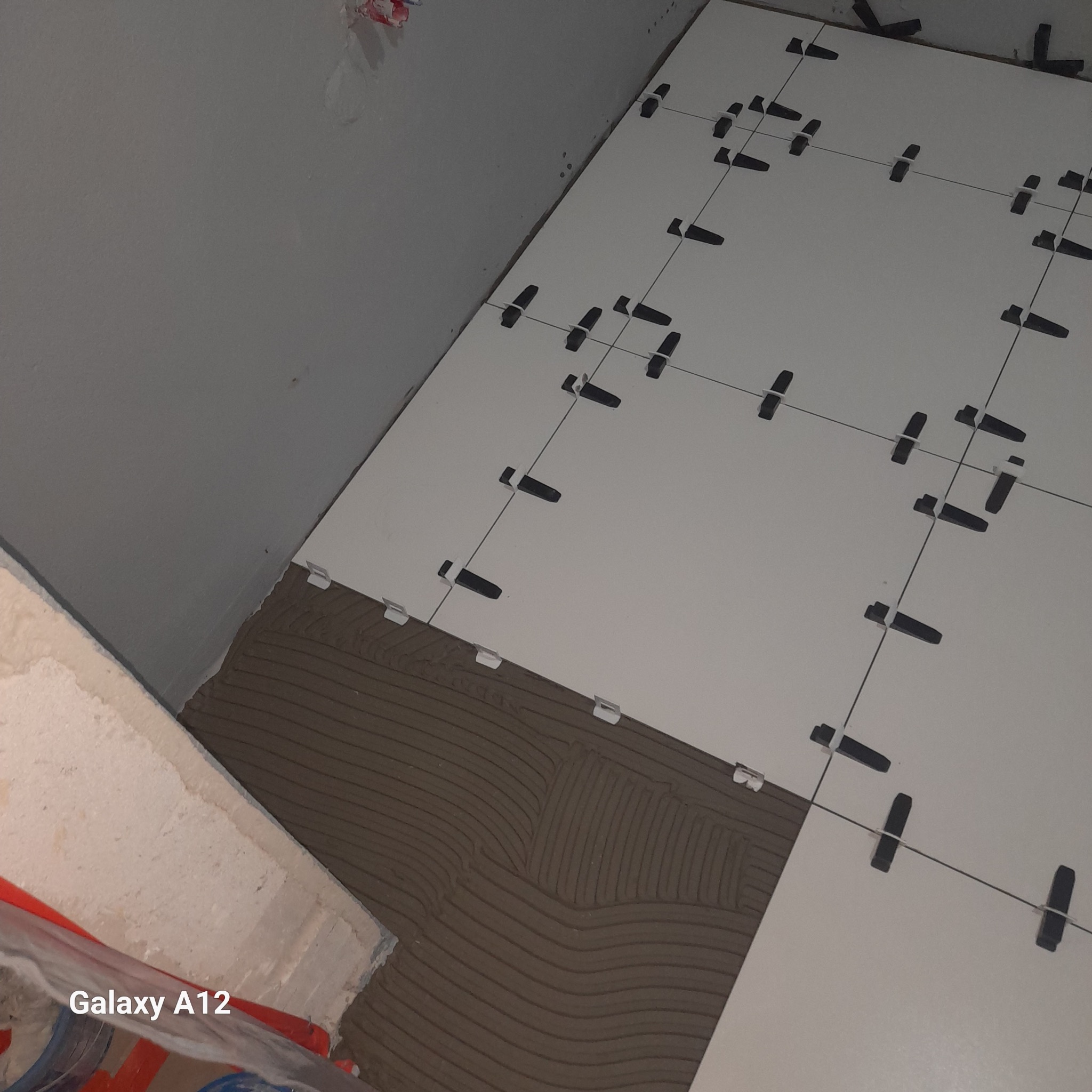 Białe płytki podłogowe w trakcie układania, z użyciem krzyżyków dystansowych i poziomowania, widoczna warstwa kleju na podłożu w rogu pomieszczenia.