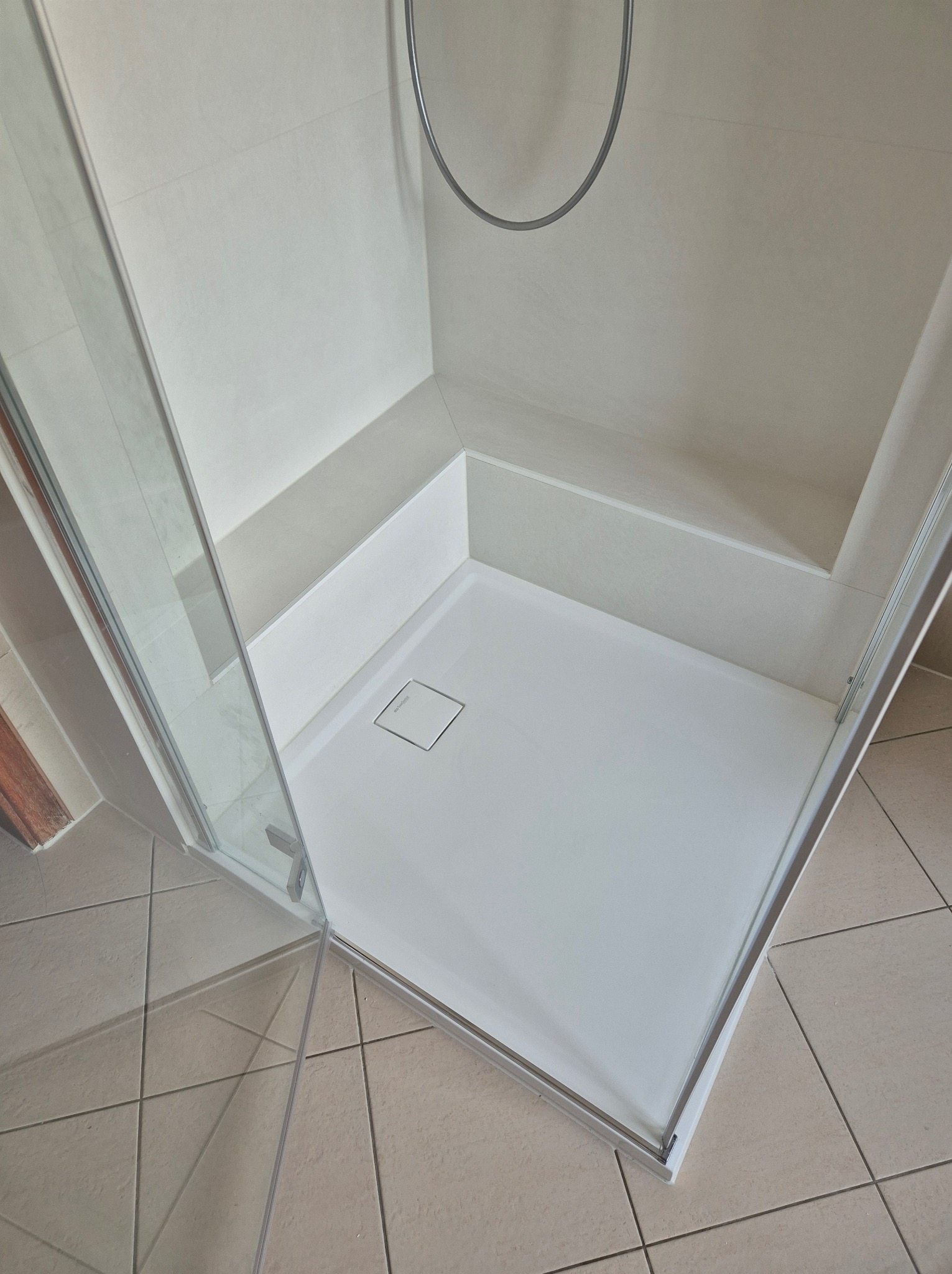 Nowoczesna kabina prysznicowa z białym brodzikiem, siedziskiem i odpływem liniowym, jasne płytki na ścianach i podłodze.