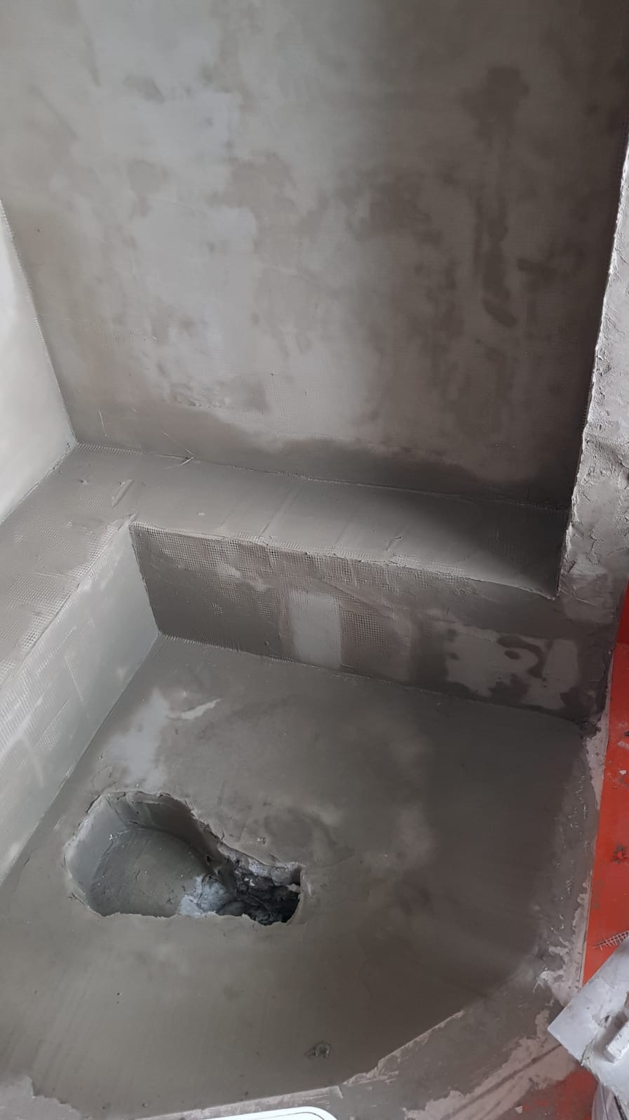 Surowa, niewykończona podłoga i ściany łazienki pokryte świeżym, szarym tynkiem. Widoczny otwór w podłodze i siatka wzmacniająca na ścianach.