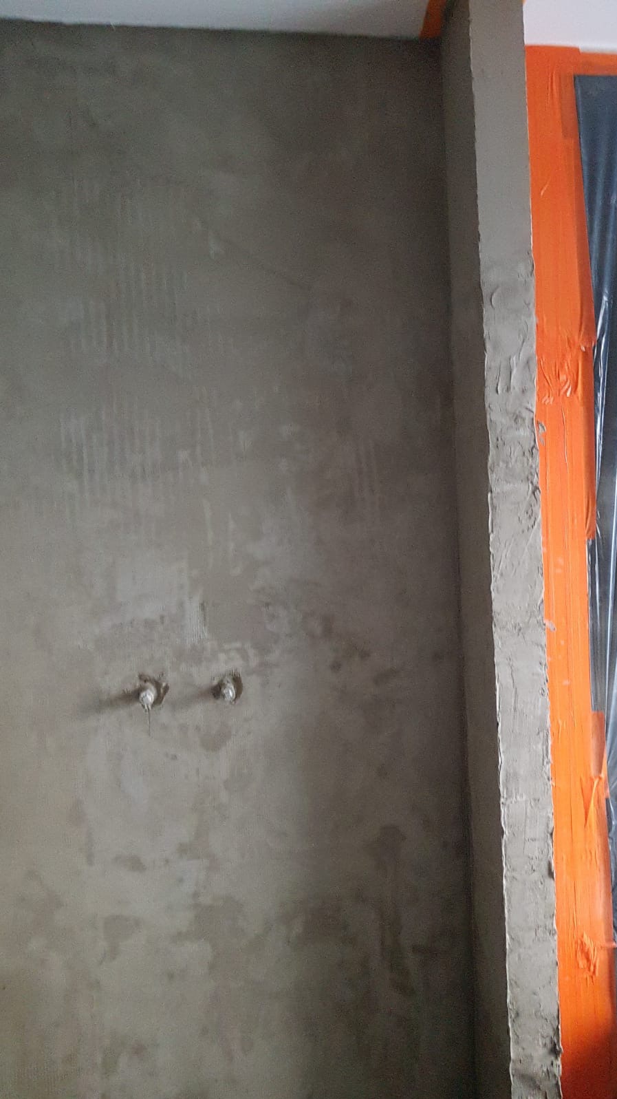 Ściana w trakcie przygotowywania do położenia płytek, widoczne wyprowadzenia hydrauliczne i zabezpieczona folią pomarańczowa ościeżnica.