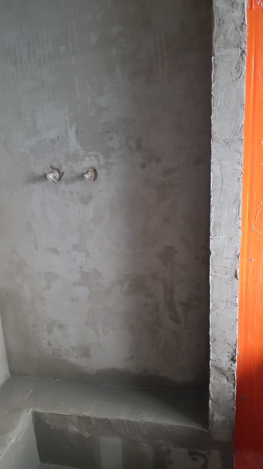 Ściana łazienki w trakcie przygotowania pod płytki, widoczne wyprowadzenia instalacji wodnej, świeżo nałożona warstwa szpachli i pomarańczowa futryna drzwi zabezpieczona folią.