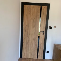 Usługi stolarsko-budowlane Kacper Głuszek - Otwarta, wewnętrzna drewniana drzwi z czarną ramą i pionowym paskiem, widoczna zamek i klamka, na podłodze panele podłogowe, w tle biała ściana z niedokończonymi gniazdkami elektrycznymi.