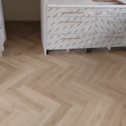 Jasne panele ułożone we wzór jodełki francuskiej, widoczne fragmenty białych szafek kuchennych z folią ochronną i złotymi uchwytami.