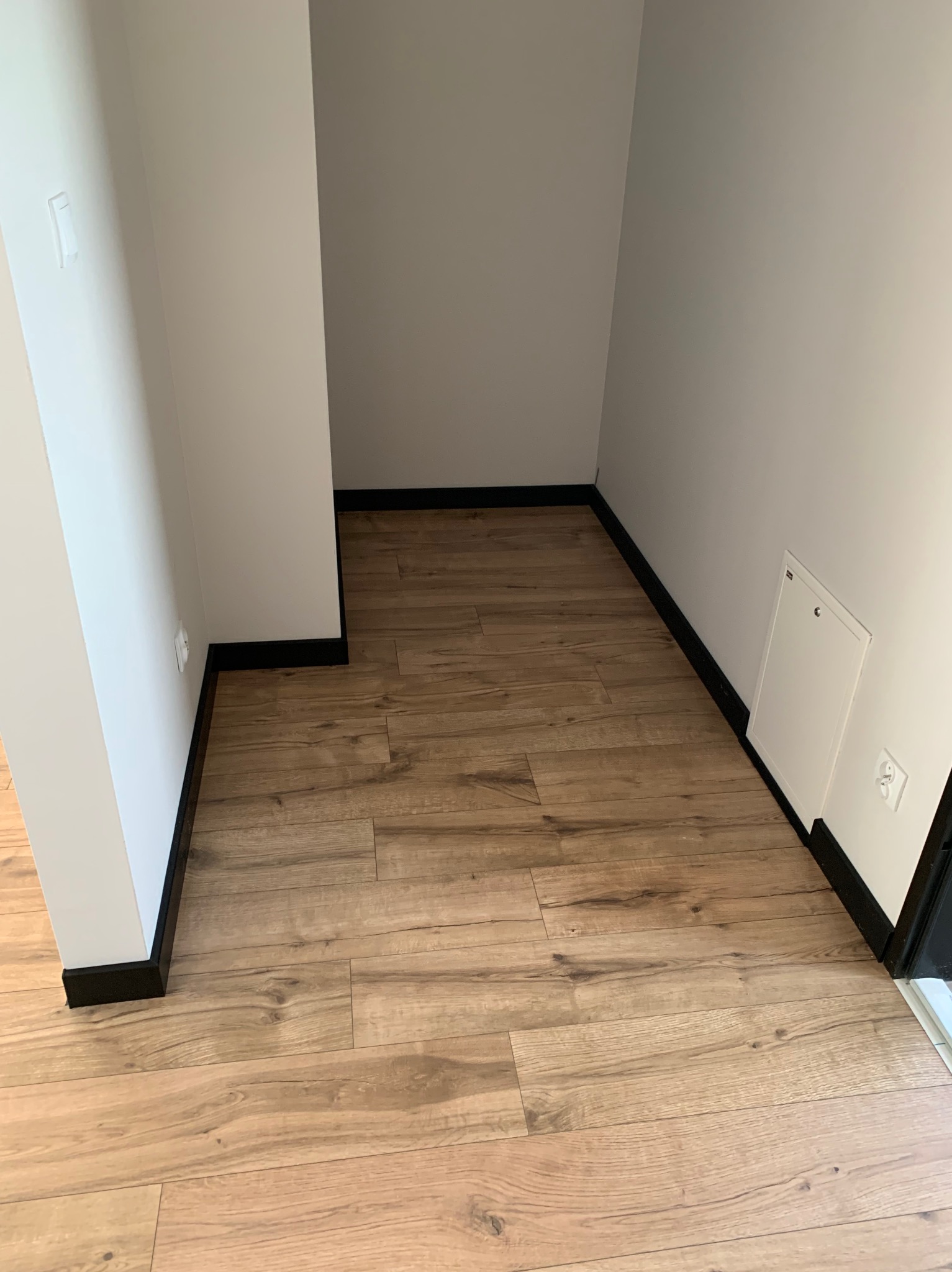 Jasnobrązowe panele podłogowe z czarnymi listwami przypodłogowymi w narożniku pomieszczenia, widoczny fragment białej ściany z gniazdkiem elektrycznym i klapką rewizyjną.