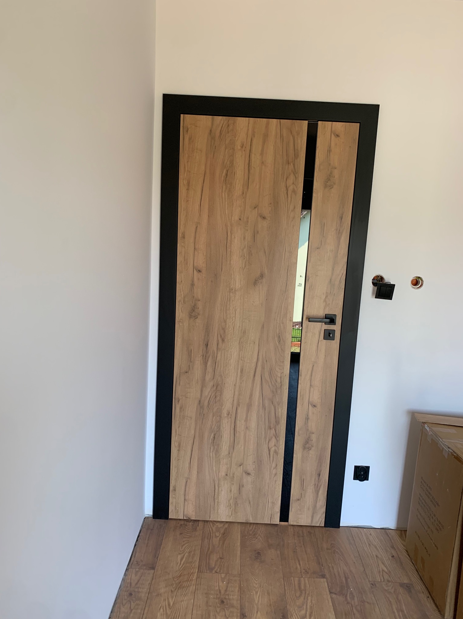 Otwarta, wewnętrzna drewniana drzwi z czarną ramą i pionowym paskiem, widoczna zamek i klamka, na podłodze panele podłogowe, w tle biała ściana z niedokończonymi gniazdkami elektrycznymi.
