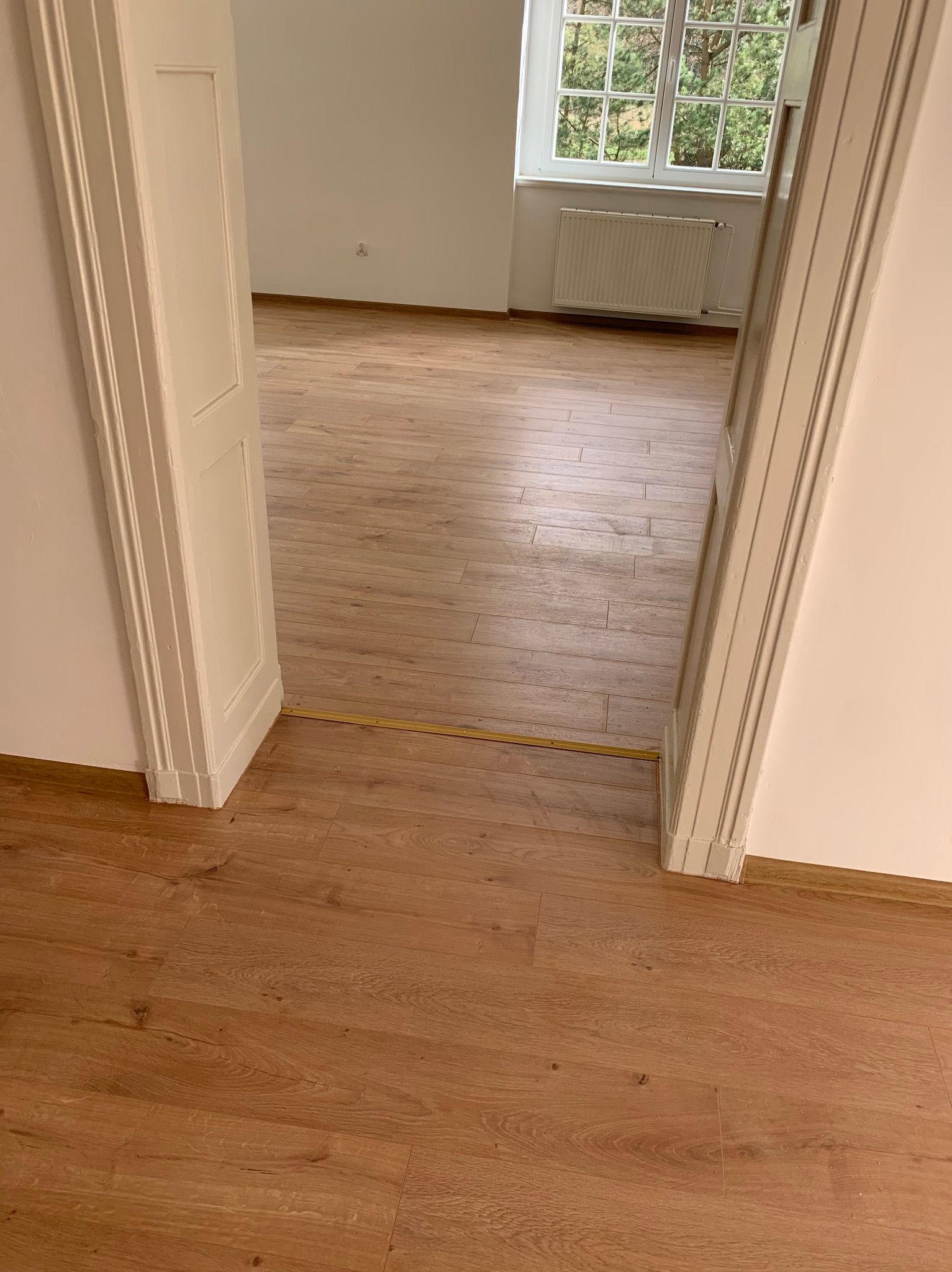 Widok z góry na świeżo ułożoną podłogę z paneli o wzorze drewna, widoczne przejście z listwą progową, białe ściany i fragment ościeżnicy drzwiowej, w tle okno z widokiem na drzewa.