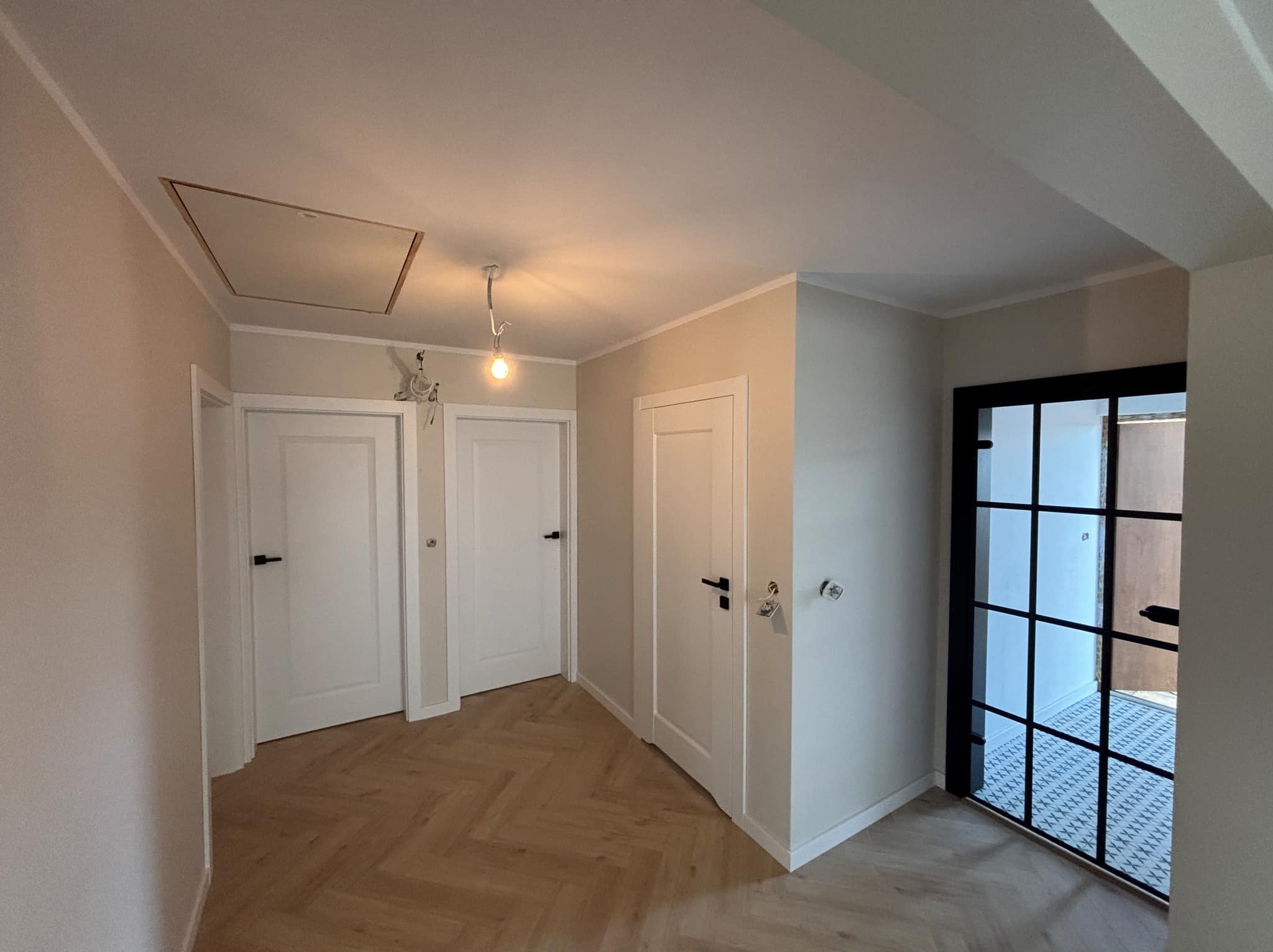 Wnętrze korytarza z parkietem ułożonym w jodełkę, trzema białymi drzwiami z czarnymi klamkami, czarną przeszkloną ścianą działową i włazem rewizyjnym w suficie.