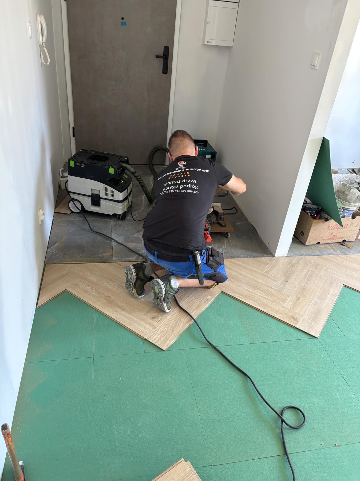 Pracownik na kolanach układa panele podłogowe w jodełkę na zielonym podkładzie, widoczny odkurzacz przemysłowy Festool i narzędzia pracy.