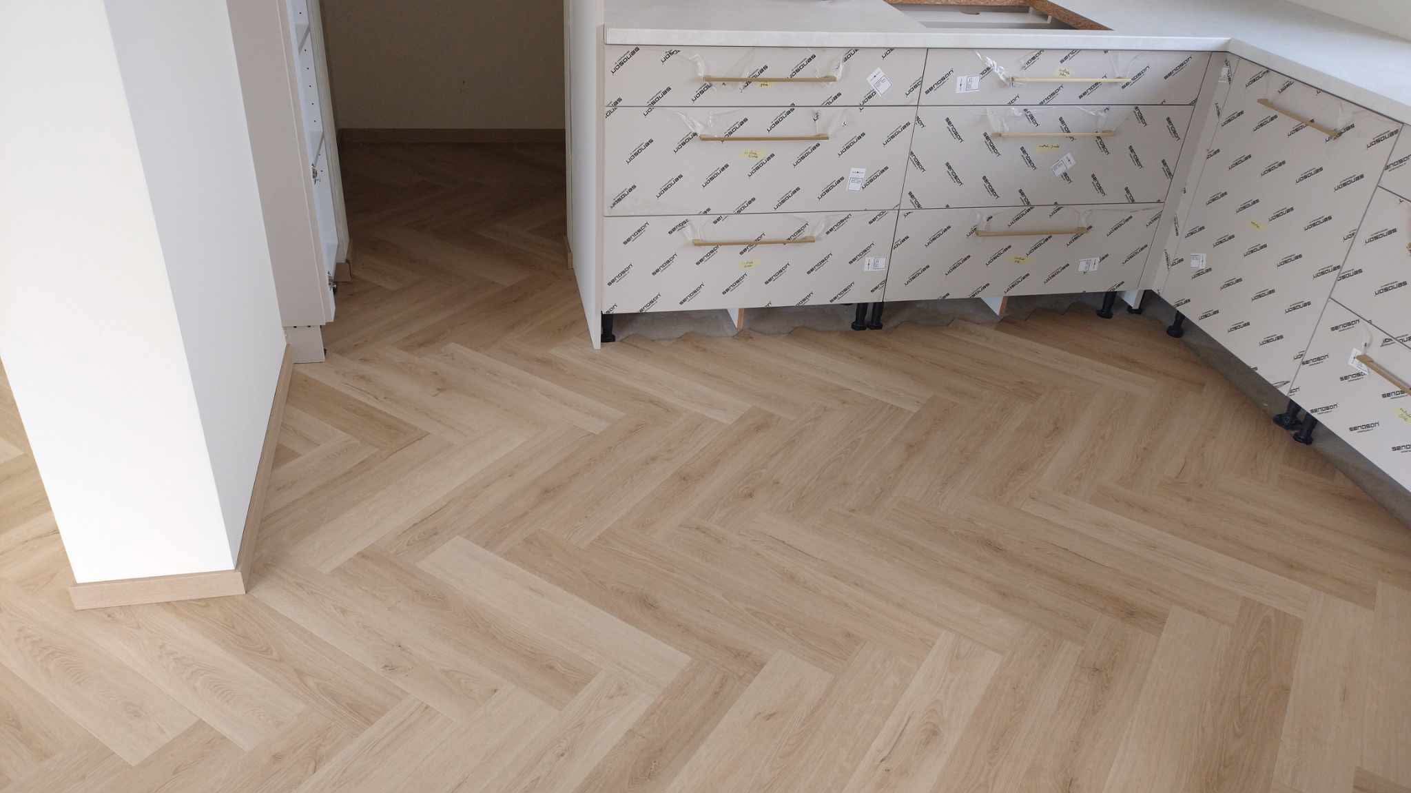 Jasne panele ułożone we wzór jodełki francuskiej, widoczne fragmenty białych szafek kuchennych z folią ochronną i złotymi uchwytami.