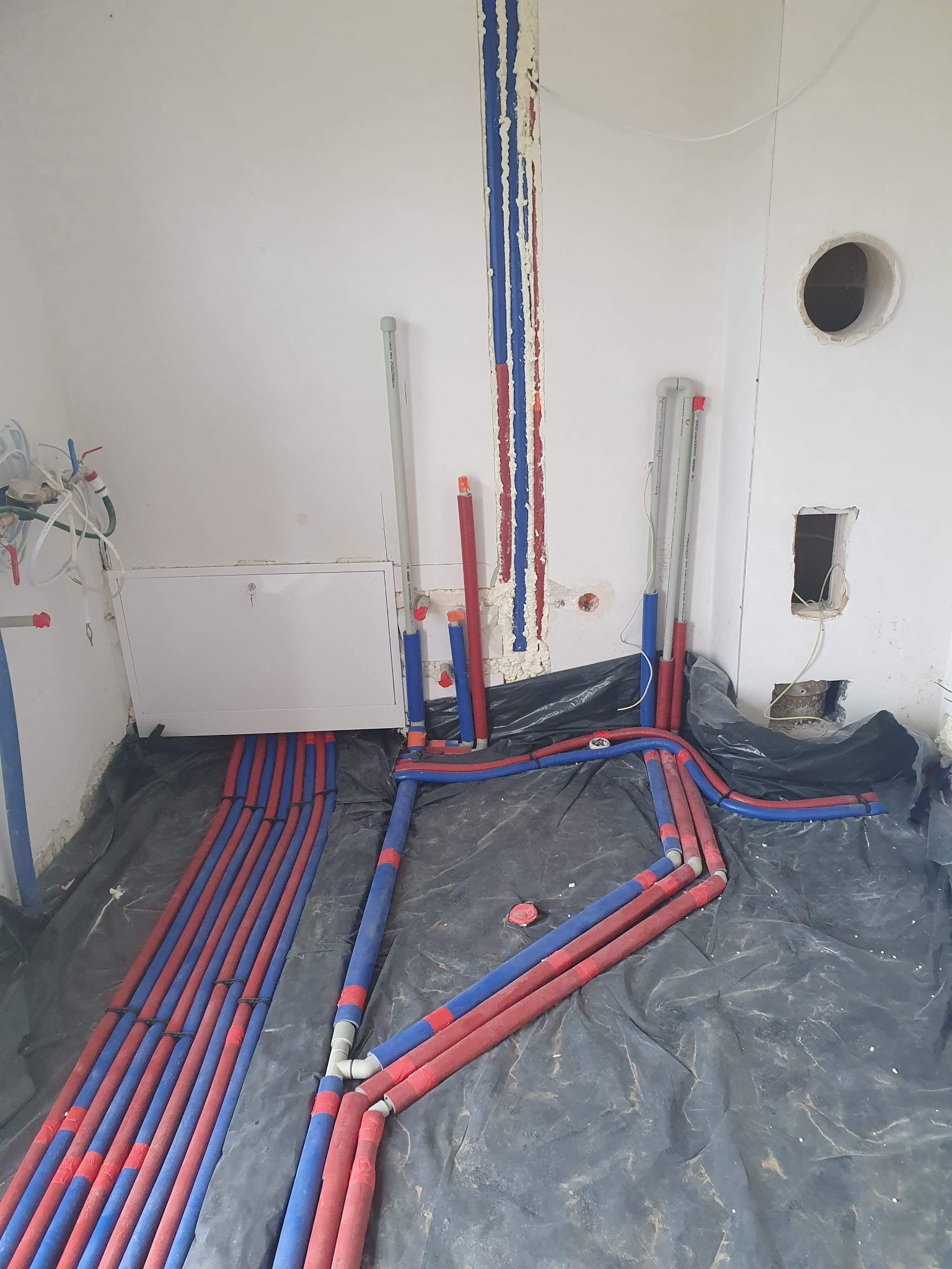 Wnętrze w trakcie prac hydraulicznych: rury grzewcze podłogowe ułożone na czarnej folii, pionowe rury przy ścianie zabezpieczone pianką montażową, widoczne otwory w ścianie.