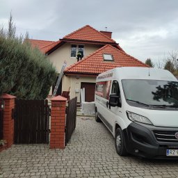 ECO-CELULOZA Piotr Kopciuch - Pracownik w zielonej kamizelce na dachu domu z czerwoną dachówką, obok drabina i biały samochód dostawczy z reklamą firmy, widok od strony brukowanej posesji.