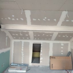 ECO-CELULOZA Piotr Kopciuch - Poddasze w trakcie wykańczania: płyty gipsowo-kartonowe na ścianach i suficie, widoczne łączenia szpachlowane, otwór do innego pomieszczenia, materiały budowlane leżące na podłodze, kartonowe...