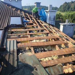 ECO-CELULOZA Piotr Kopciuch - Prace na dachu: demontaż starego poszycia i ocieplenia, widoczne drewniane elementy konstrukcji, resztki izolacji i pracownik w zielonej koszulce z workiem na odpady.
