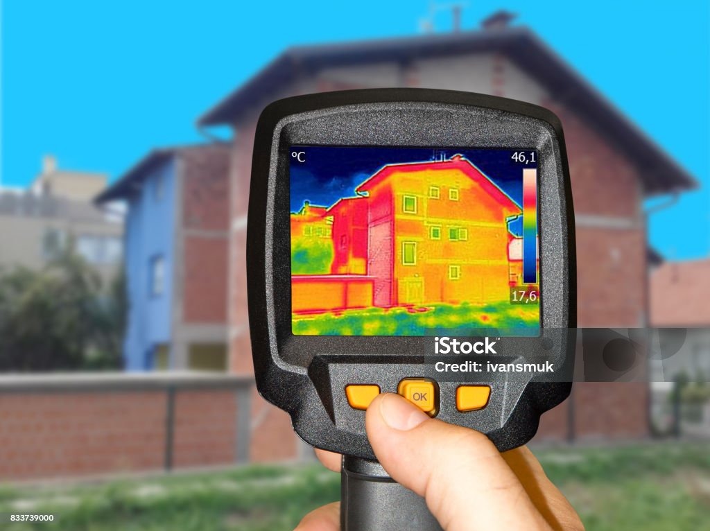 Termowizja budynku mieszkalnego na ekranie kamery termowizyjnej trzymanej w dłoni, z widocznymi wskazaniami temperatury.