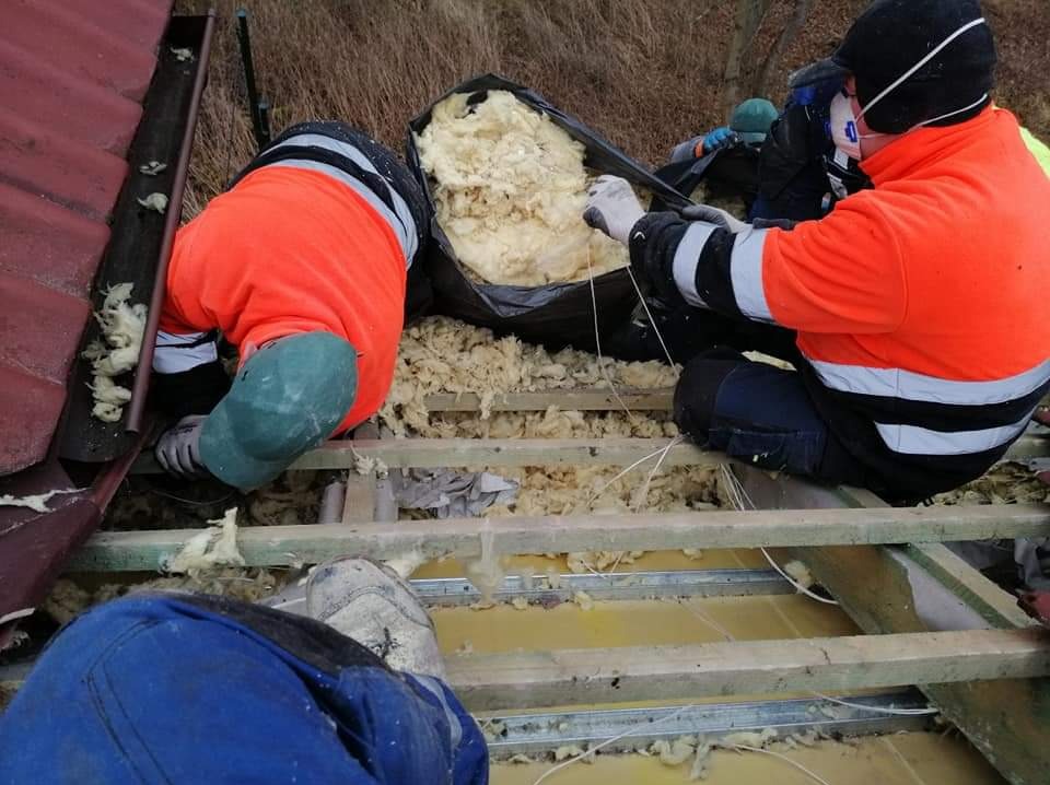 Pracownicy w odblaskowych ubraniach układają izolację z wełny mineralnej na drewnianej konstrukcji poddasza, widoczny fragment dachu z rynną.