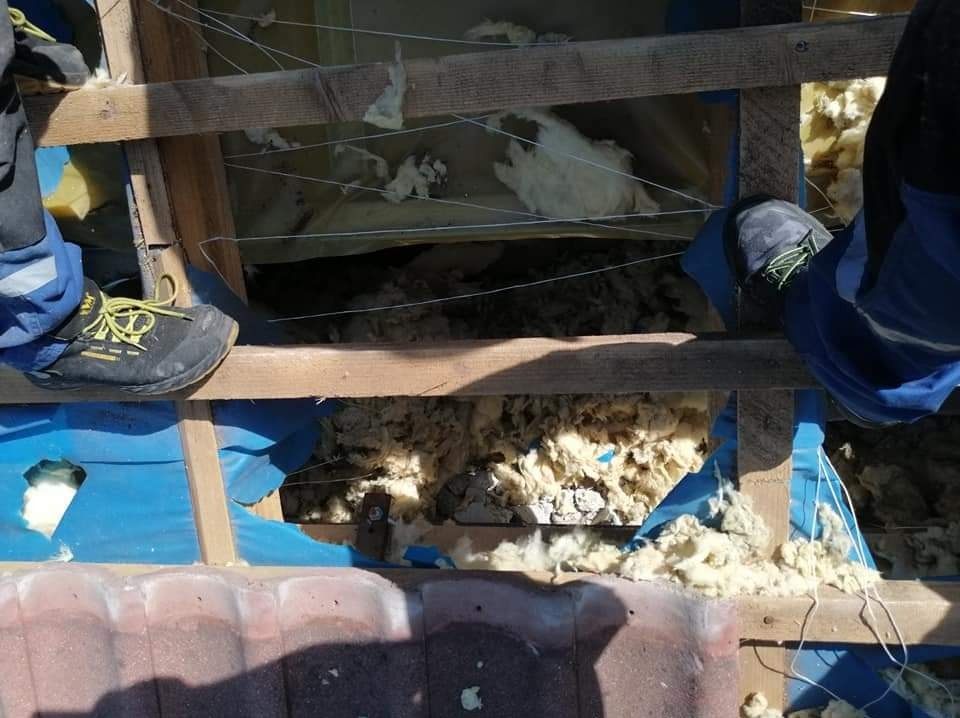 Montaż izolacji z wełny mineralnej na poddaszu, widoczne drewniane krokwie i fragment dachu z dachówkami, fragment odzieży roboczej montera.