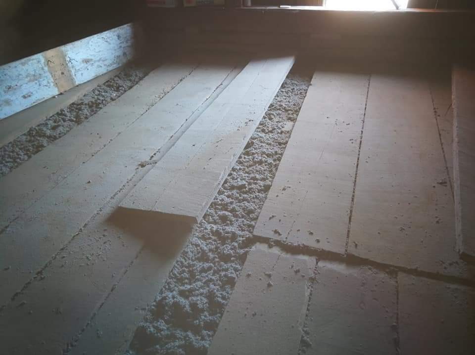 Poddasze w trakcie izolacji: widoczne deski podłogowe z przestrzeniami wypełnionymi wełną izolacyjną, skierowane w stronę okna z jasnym światłem.