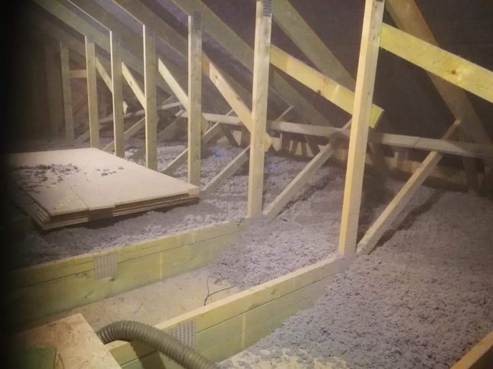 Poddasze w trakcie izolacji: drewniana konstrukcja więźby dachowej, warstwa szarej wełny mineralnej i stos płyt OSB, widoczny wąż od maszyny do wdmuchiwania izolacji.