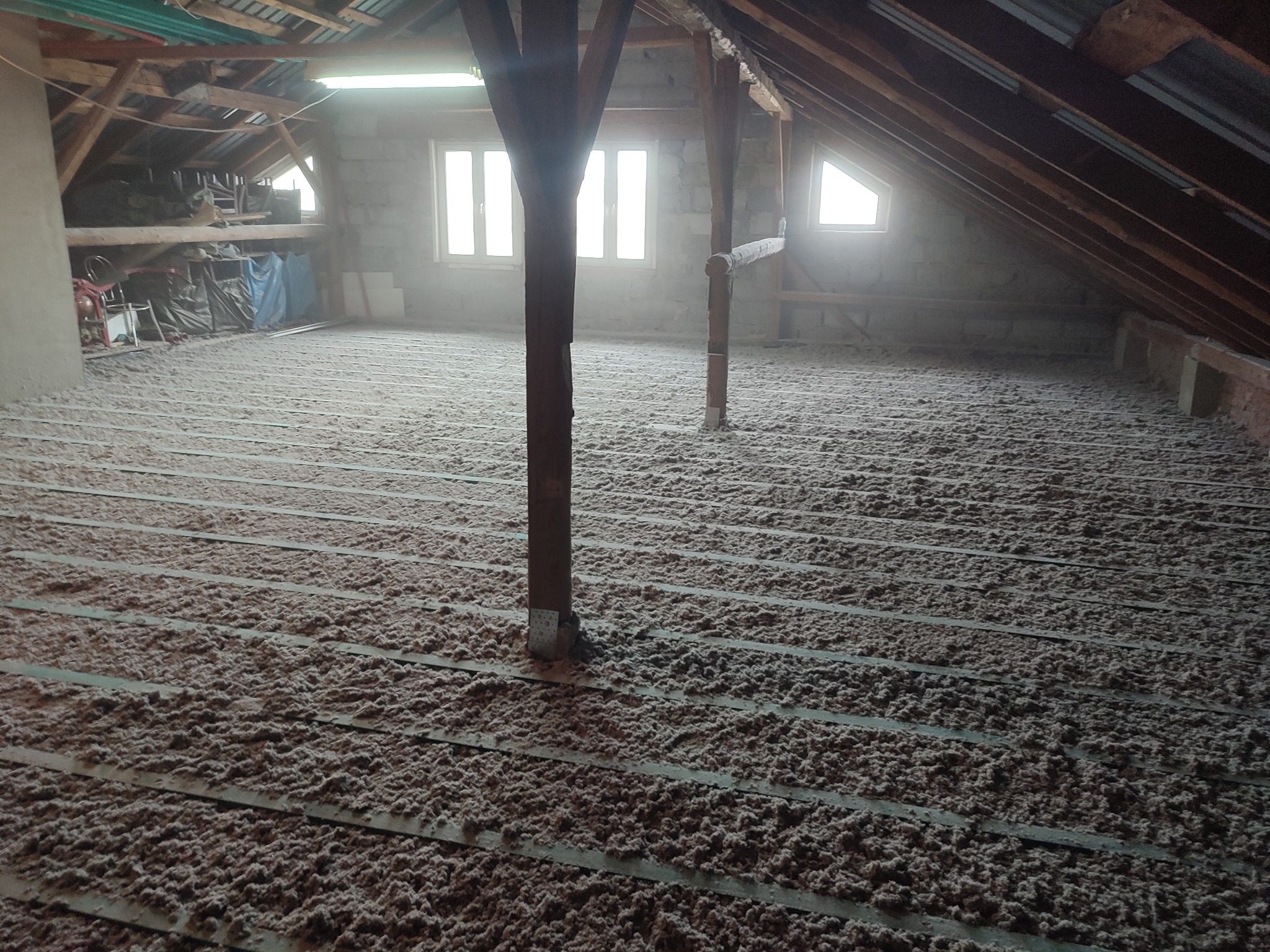 Poddasze z widoczną izolacją z wełny mineralnej ułożoną między legarami, drewniane słupy podtrzymujące konstrukcję dachu, okna dachowe w tle.