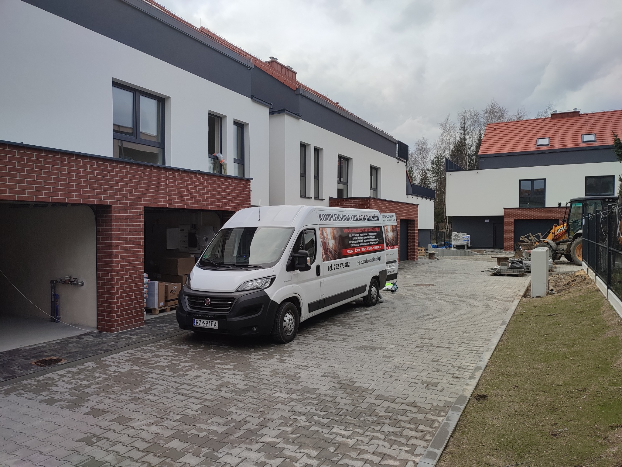 Biały bus firmy specjalizującej się w izolacji dachów zaparkowany na brukowanym podjeździe przed szeregiem nowoczesnych domów w trakcie prac budowlanych, widoczny pracownik przy oknie jednego z domów.