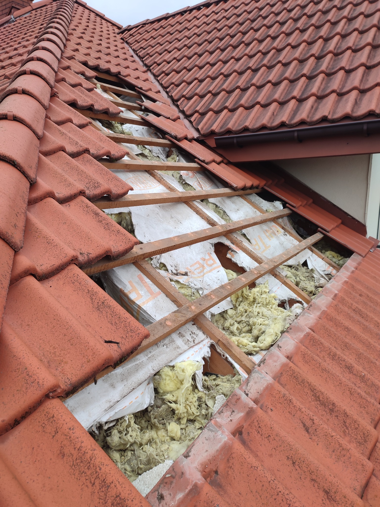Fragment dachu z usuniętymi dachówkami, ukazujący drewnianą konstrukcję i fragmenty izolacji z wełny mineralnej oraz białej folii paroizolacyjnej.