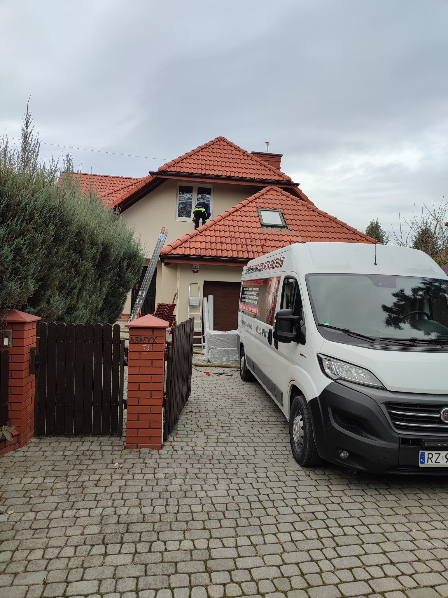 Pracownik w zielonej kamizelce na dachu domu z czerwoną dachówką, obok drabina i biały samochód dostawczy z reklamą firmy, widok od strony brukowanej posesji.