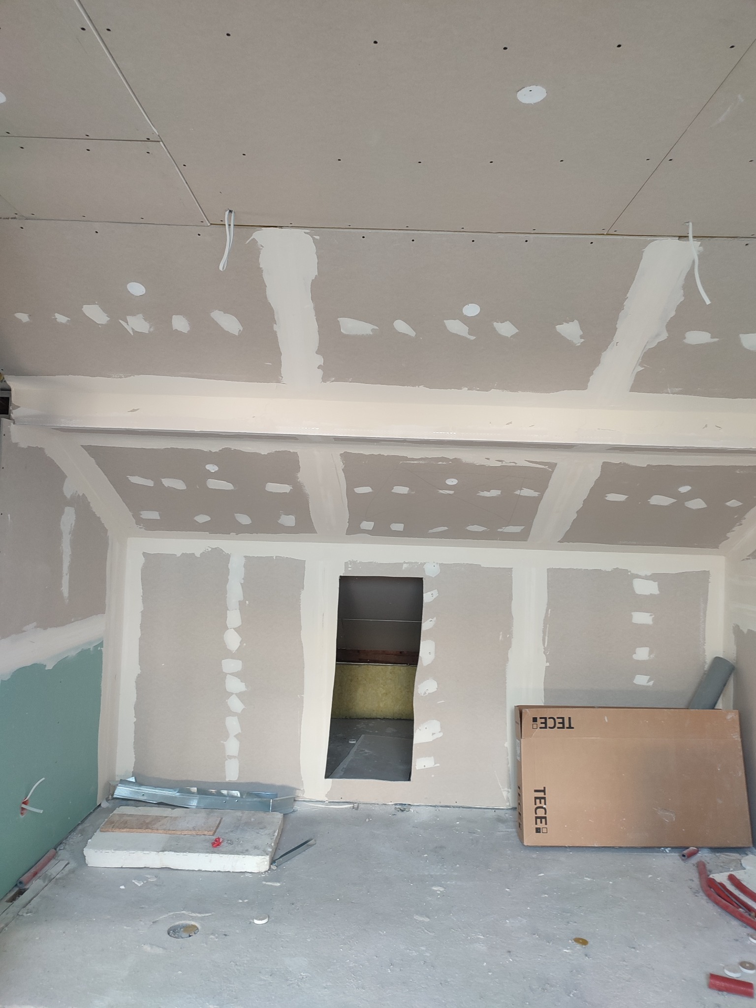 Poddasze w trakcie wykańczania: płyty gipsowo-kartonowe na ścianach i suficie, widoczne łączenia szpachlowane, otwór do innego pomieszczenia, materiały budowlane leżące na podłodze, kartonowe...