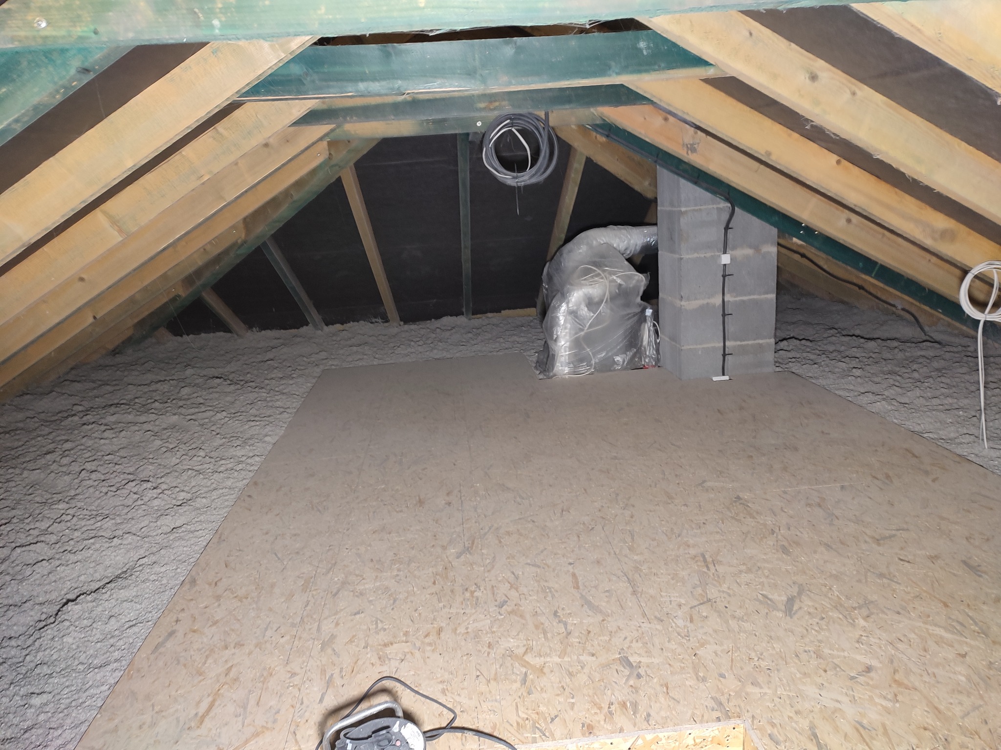 Poddasze z drewnianą konstrukcją dachu, częściowo wyłożone płytami OSB i pokryte szarą wełną mineralną, widoczny komin z pustaków i zwisające przewody elektryczne.