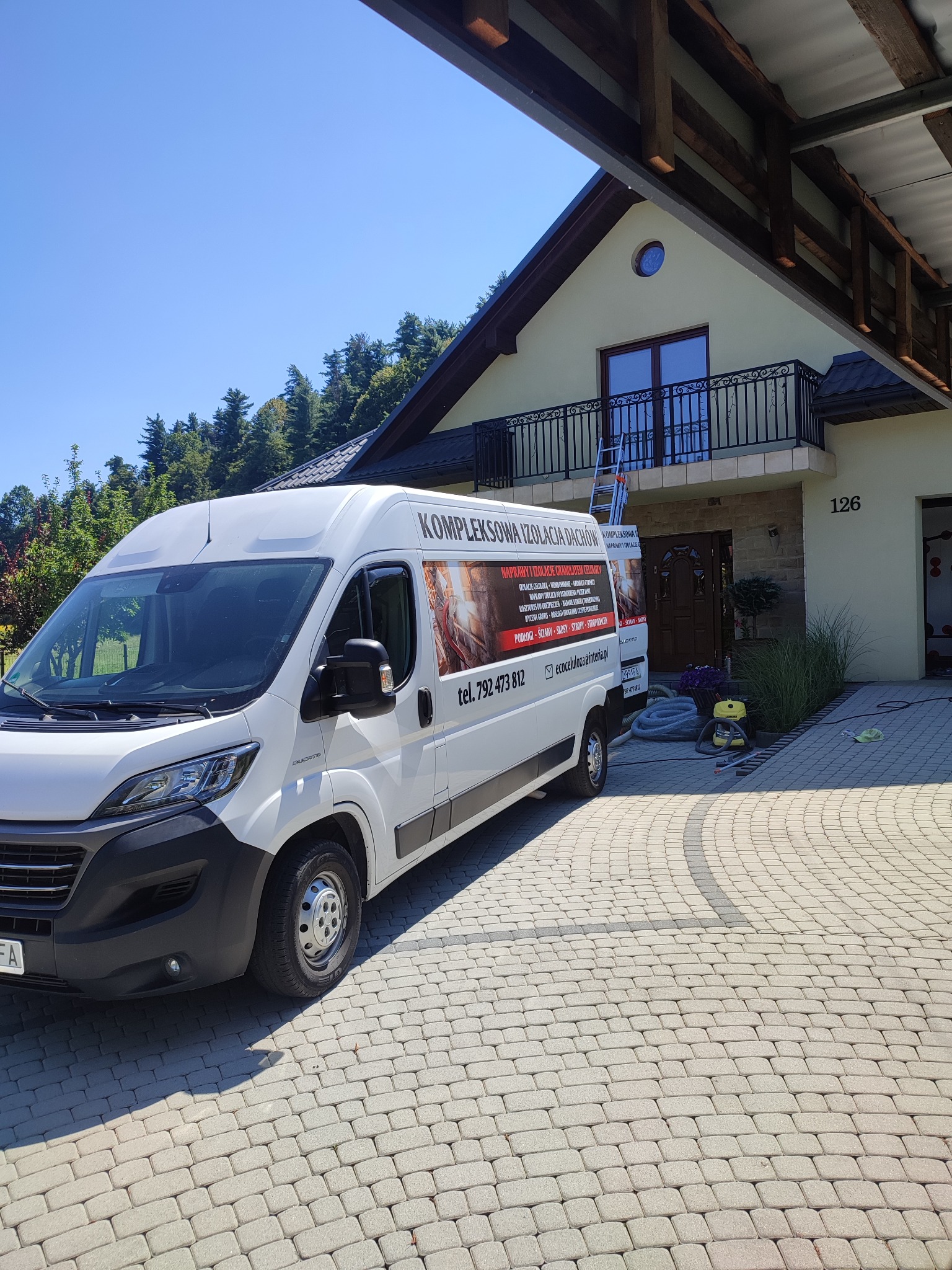 Biały samochód dostawczy firmy specjalizującej się w izolacjach dachowych, zaparkowany przed domem z balkonem i numerem 126, widoczna drabina oparta o balkon, węże i urządzenie do natrysku izolacji.