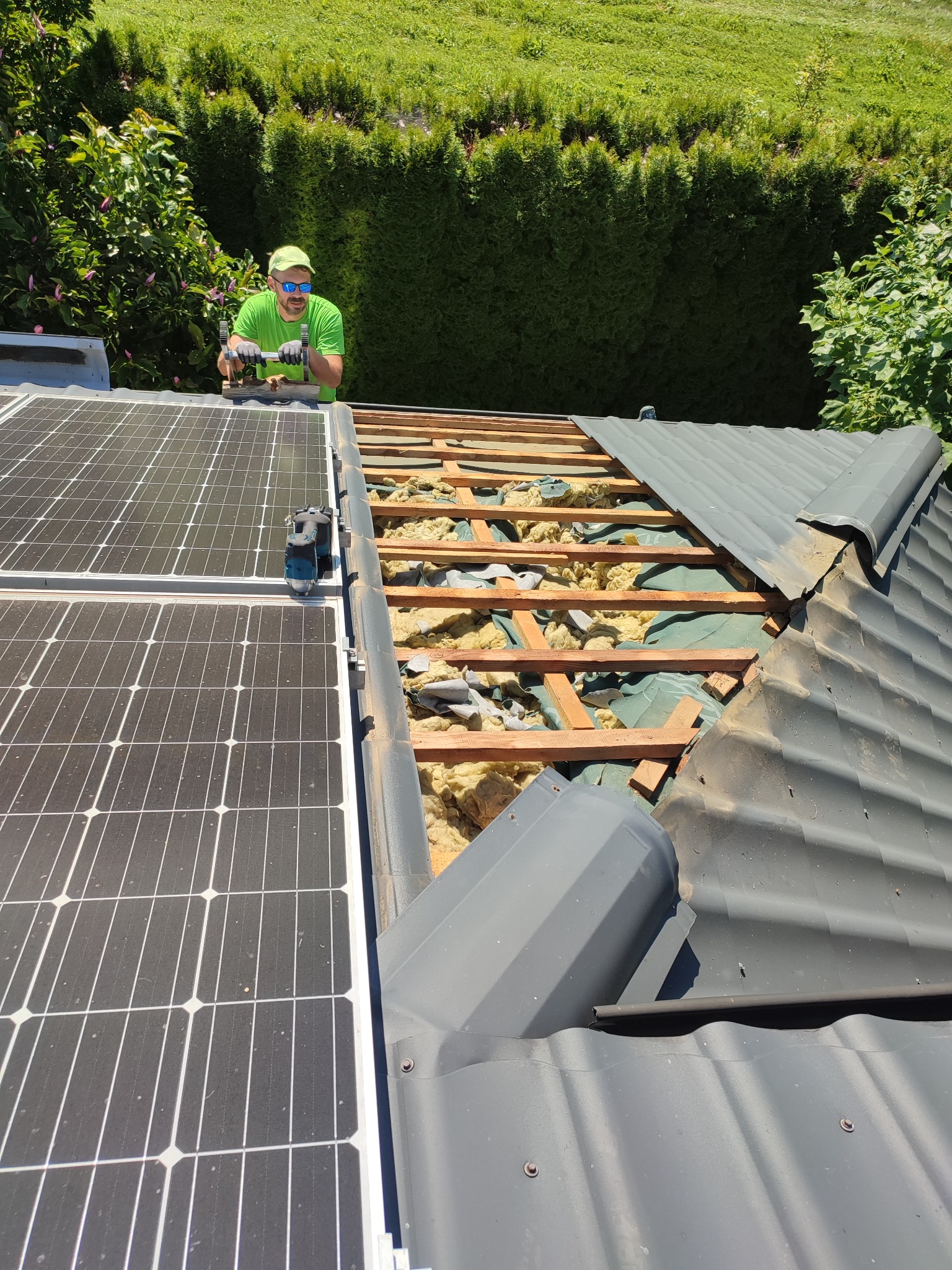 Pracownik w zielonej koszulce i okularach, z wiertarką na dachu z częściowo zdjętym pokryciem, widoczne panele słoneczne i izolacja z wełny mineralnej.