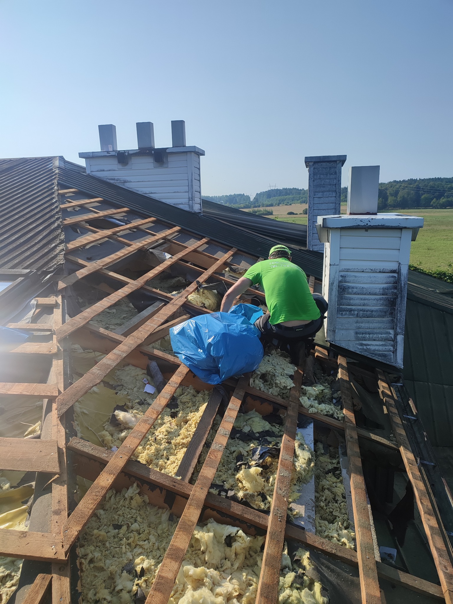 Pracownik w zielonej koszulce usuwa starą izolację z poddasza, widoczne drewniane elementy konstrukcji dachu i białe kominy na tle zielonych pól.