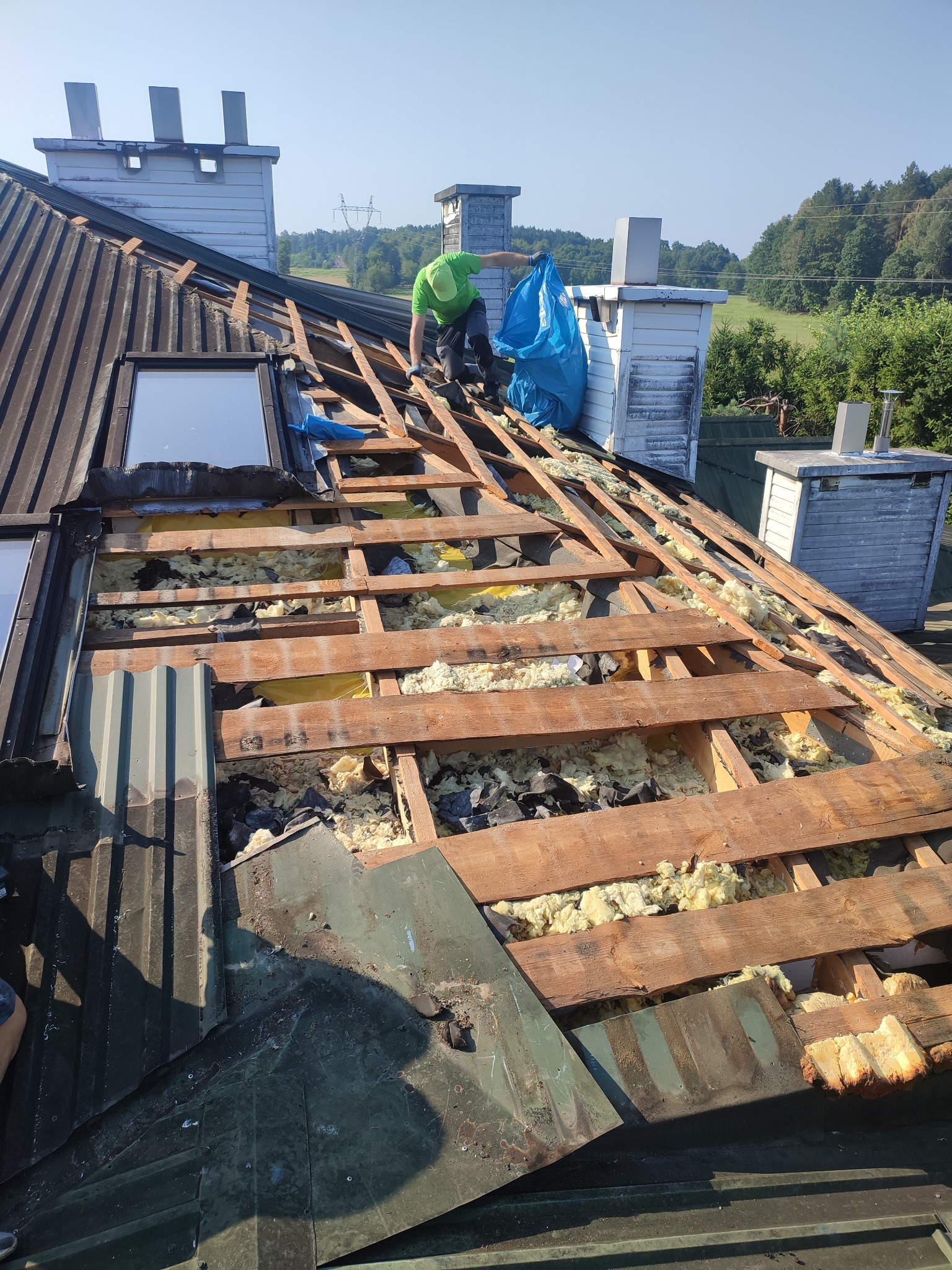 Prace na dachu: demontaż starego poszycia i ocieplenia, widoczne drewniane elementy konstrukcji, resztki izolacji i pracownik w zielonej koszulce z workiem na odpady.