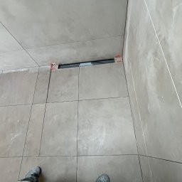 Jan Antoszczyk - Beżowe płytki podłogowe i ścienne w trakcie montażu, widoczne elementy dystansowe. U dołu kadru fragment obuwia osoby wykonującej prace.