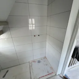 Białe, błyszczące płytki w kabinie prysznicowej z widocznymi przyłączami wodnymi. Na podłodze mata ochronna. Narzędzie glazurnicze leży obok.