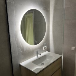 Jan Antoszczyk - Szara łazienka z okrągłym lustrem LED, białą umywalką na drewnianej szafce i szklaną kabiną prysznicową. Minimalistyczny design, płytki imitujące kamień.