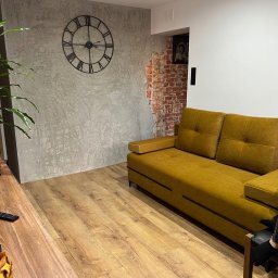 Jan Antoszczyk - Nowoczesne wykończenie wnętrza: szara ściana z efektem betonu, zegar rzymski i fragment odsłoniętej cegły. Ustawiona na parkiecie musztardowa kanapa.