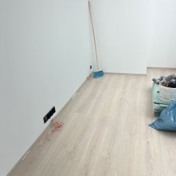 Jan Antoszczyk - Pokój po remoncie: białe ściany, jasna podłoga, listwy przypodłogowe. Widoczna szczotka oparta o ścianę, gniazdka elektryczne i narzędzia w koszu.